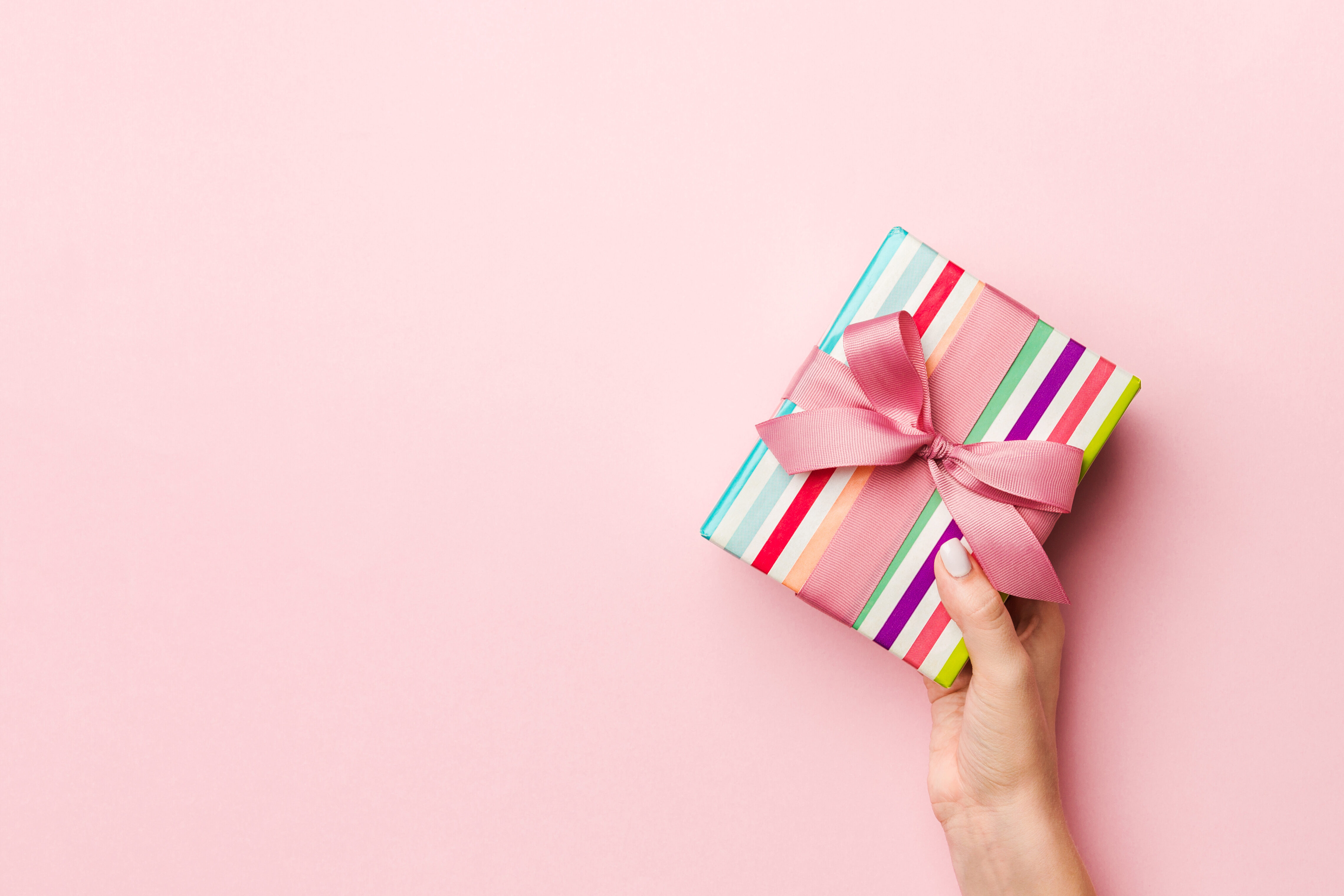 Female's Hands Holding Striped Gift Box With Colored Ribbon On Living Coral Background. Christmas Concept Or Other Holiday Handmade Present Box, Concept Top View With Copy Space